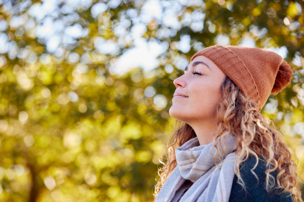 A woman in a hat and scarf pausing outdoors among autumn trees.